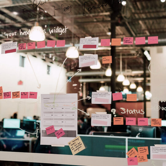 Photo vitre sur laquelle sont disposés des post-it avec des inscriptions avec un open space de bureaux en arrière plan pour illustrer la Demand Path Optimisation