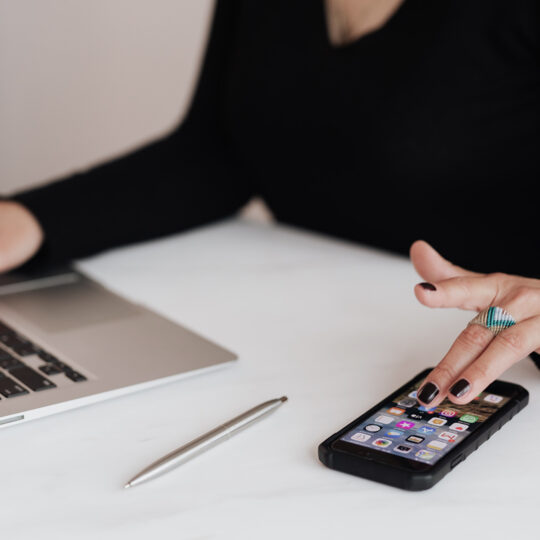 Foto de una persona con un suéter negro sentada detrás de un escritorio blanco usando una computadora portátil mientras abre una aplicación móvil en su teléfono