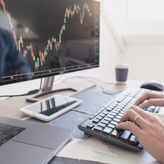 Foto de una persona sentada detrás de un escritorio escribiendo en el teclado de una computadora y viendo los cambios de precios en las pantallas